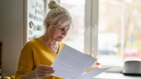 Getty Images Woman looks at paperwork in a kitchen with a fridge-freezer and a toaster behind her.