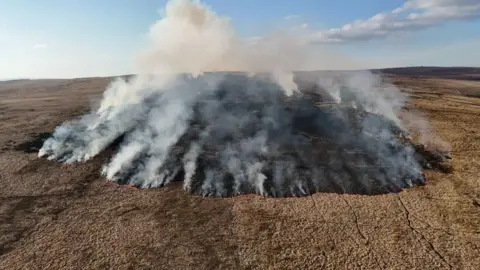 Eddy Blanche An aerial photo showing a mountainside with a ring of smoke and blackened land in the centre as a grassfire fire burns across the mountain.