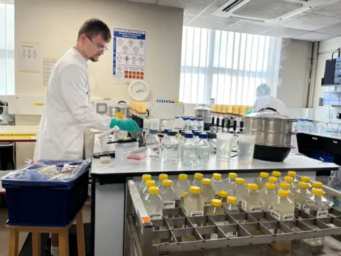 Jonah Fisher/BBC A man clad in a white jacket and wearing glasses conducts water testing at the Starcross laboratory in Devon.
