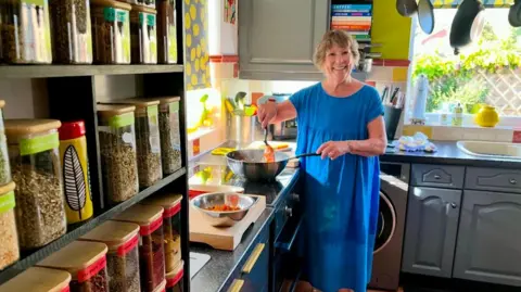 Positive Community Action A woman in a blue dress cooks a meal in a pan in a tidy kitchen and smiles at the camera. In the foreground are several herbs and spices.