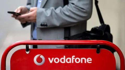 Getty Images A man wearing a suit and holding his phone stands in front of a Vodafone sign.