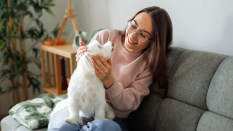 Getty Images A young woman sits at home on a grey sofa cuddling her small white dog.