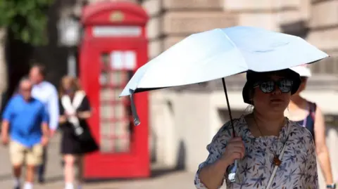 EPA A woman, wearing a sunhat and sunglasses, holds an umbrella to shade herself as she walks along a street in London. There is a red telephone box behind her as well as a few other pedestrians.